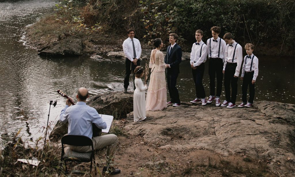 A small intimate micro wedding next to a river. In this photo is a small wedding taking place with a bride, groom, pastor, 5 children, and a man playing an acoustic guitar.