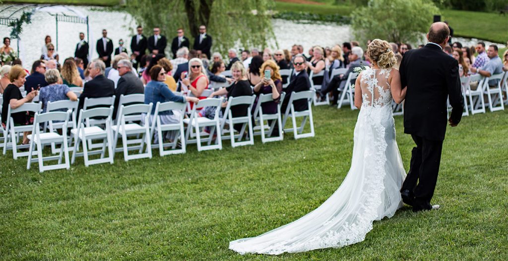 A bride walking down the aisle to a modern instrumental song on her wedding day. The photo is taken from the back of the aisle.