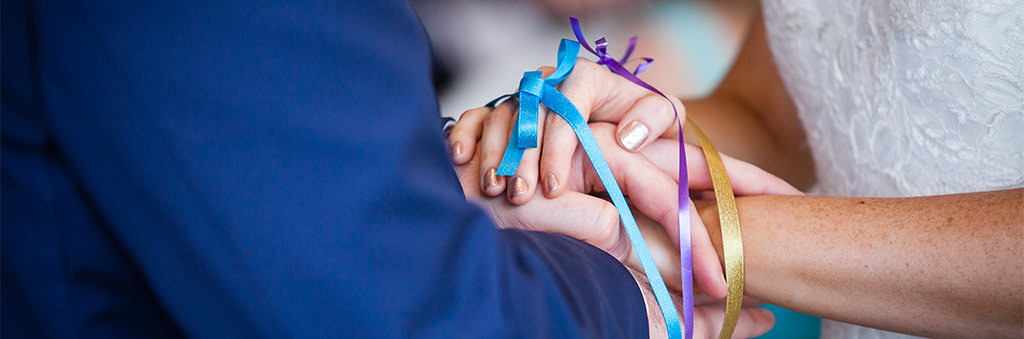 A close up of a handfasting unity ceremony at a wedding. The hands are wrapped in blue and purple ribbon