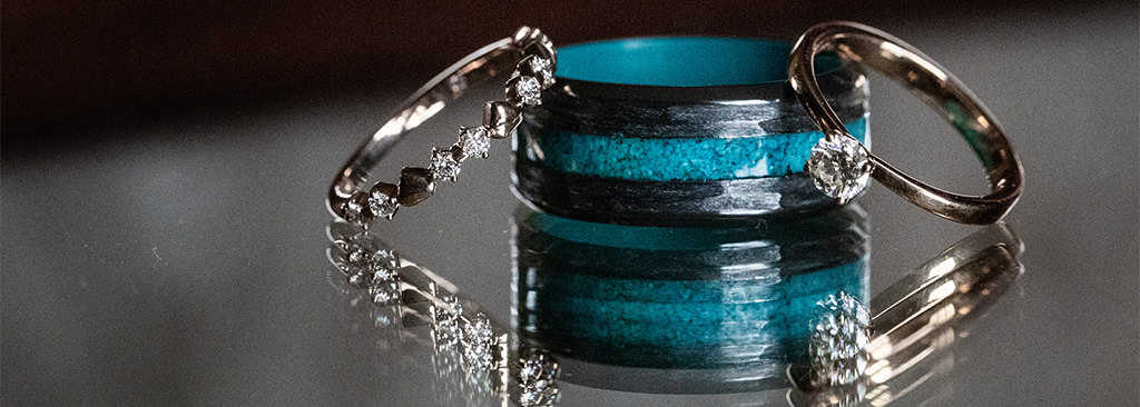 A black and turquoise wedding band for a man sitting a table with the bride's ring and engagement ring. This is for the article called Best Material for Men’s Wedding Bands