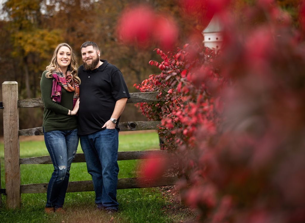 A couples posing for their engagement photos near a bright red bush in the fall