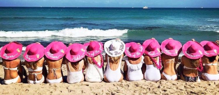 This is the cover photo for the article called beast beach bachelorette party destinations. This photo shows a group of girls with pink hats sitting on the beach with the bride in the middle wearing a white hat that says bride.