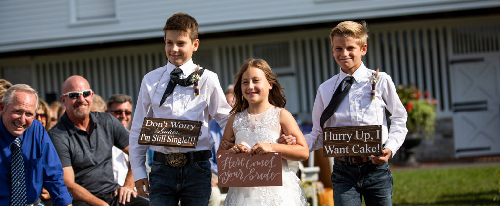 Two rings bearers and a flower girl walking down the aisle at a wedding ceremony holding fun signs