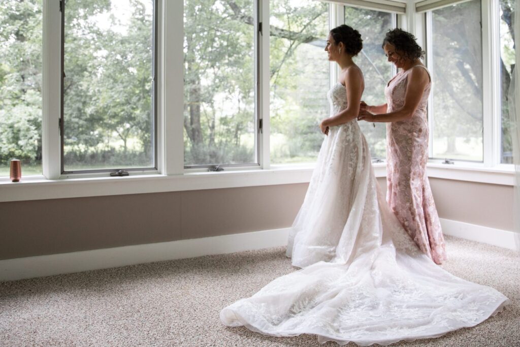 A mother of the bride zipping up the bride's wedding dress in front of a big window
