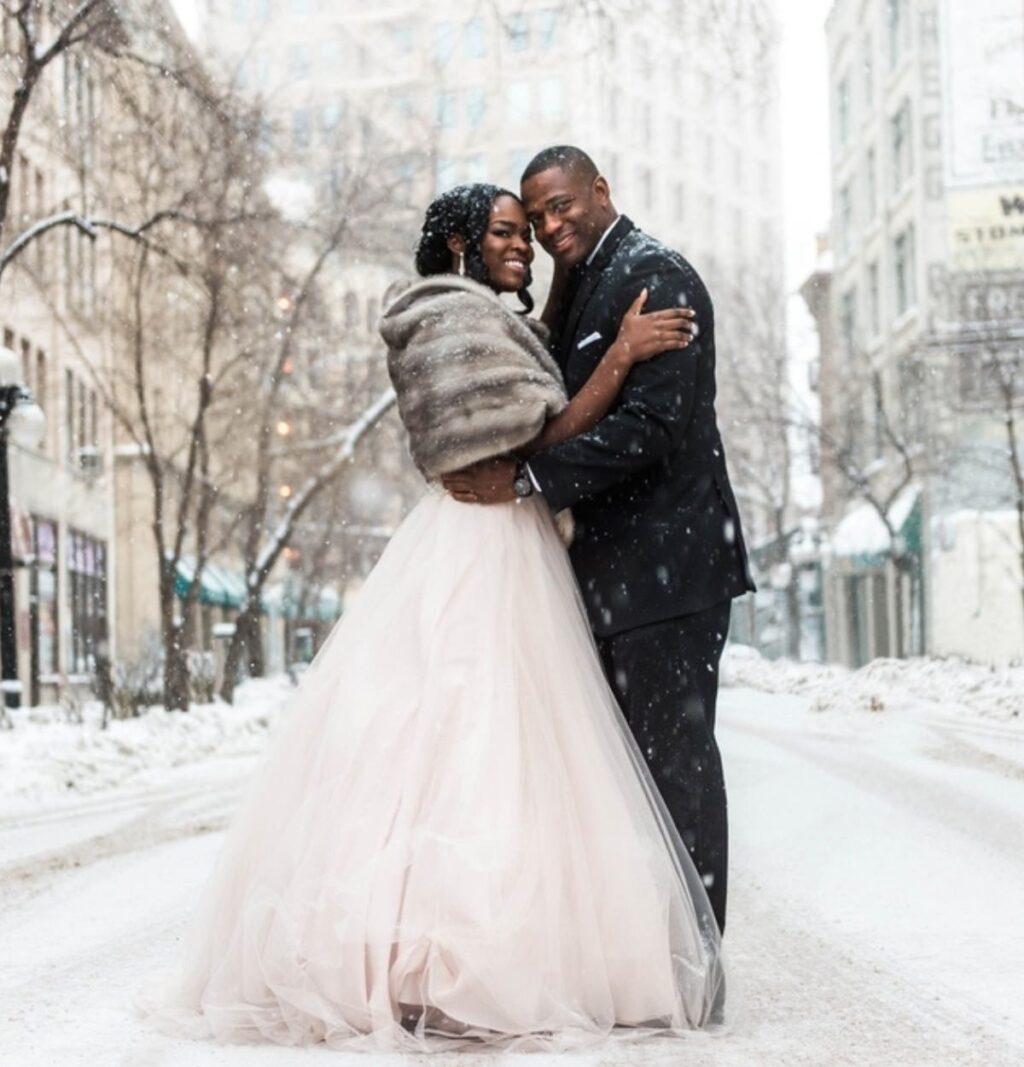 A bride and groom posing in the middle of a snow filled street in the city. The bride is wearing her wedding dress with a faux fur wrap.