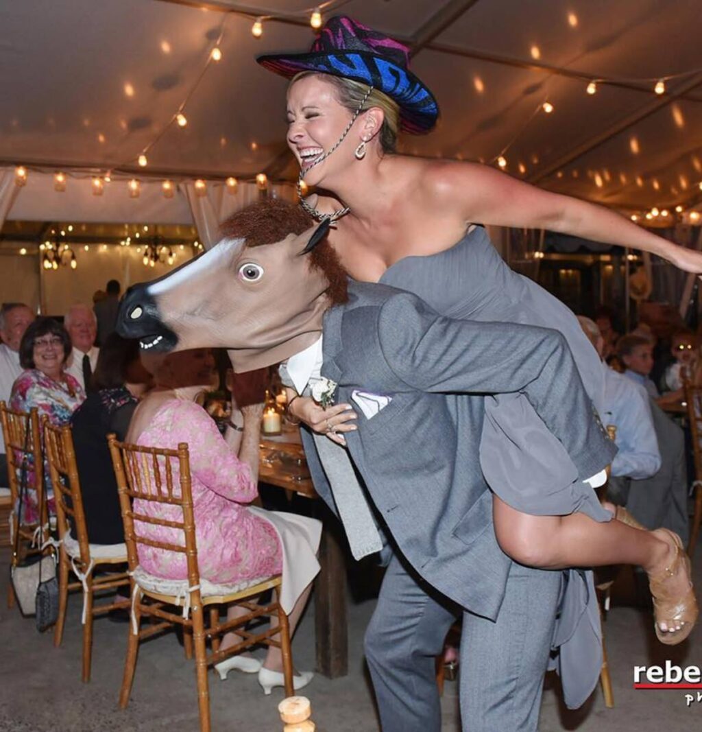 A funny photo of a bridesmaid riding on the back of a groomsmen as they enter the wedding reception. The groomsmen is wearing a rubber horse mask.