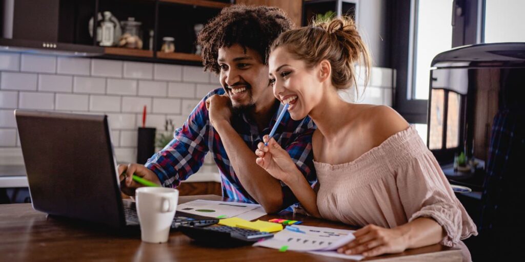 A couple sitting down to figure out their wedding budget. They are smiling looking at a computer. This is for the article called how to plan a wedding in 2025