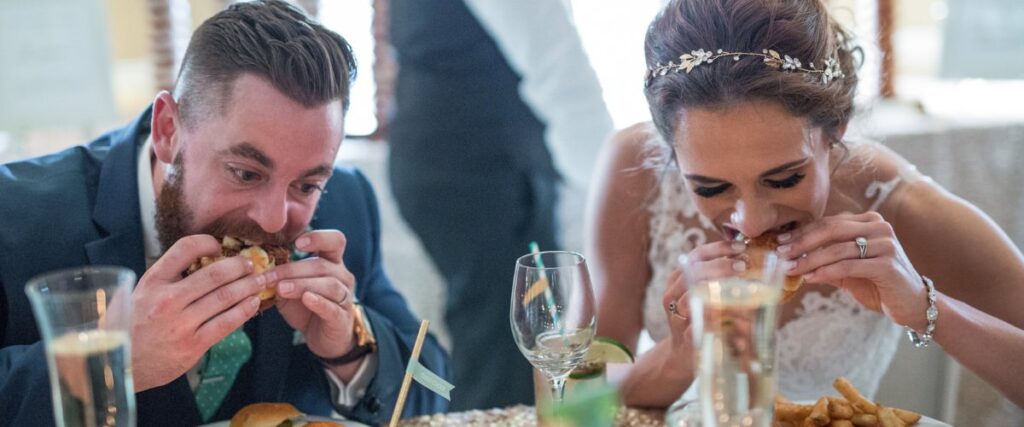 A bride and groom eating at their wedding. Taking big bites of burgers
