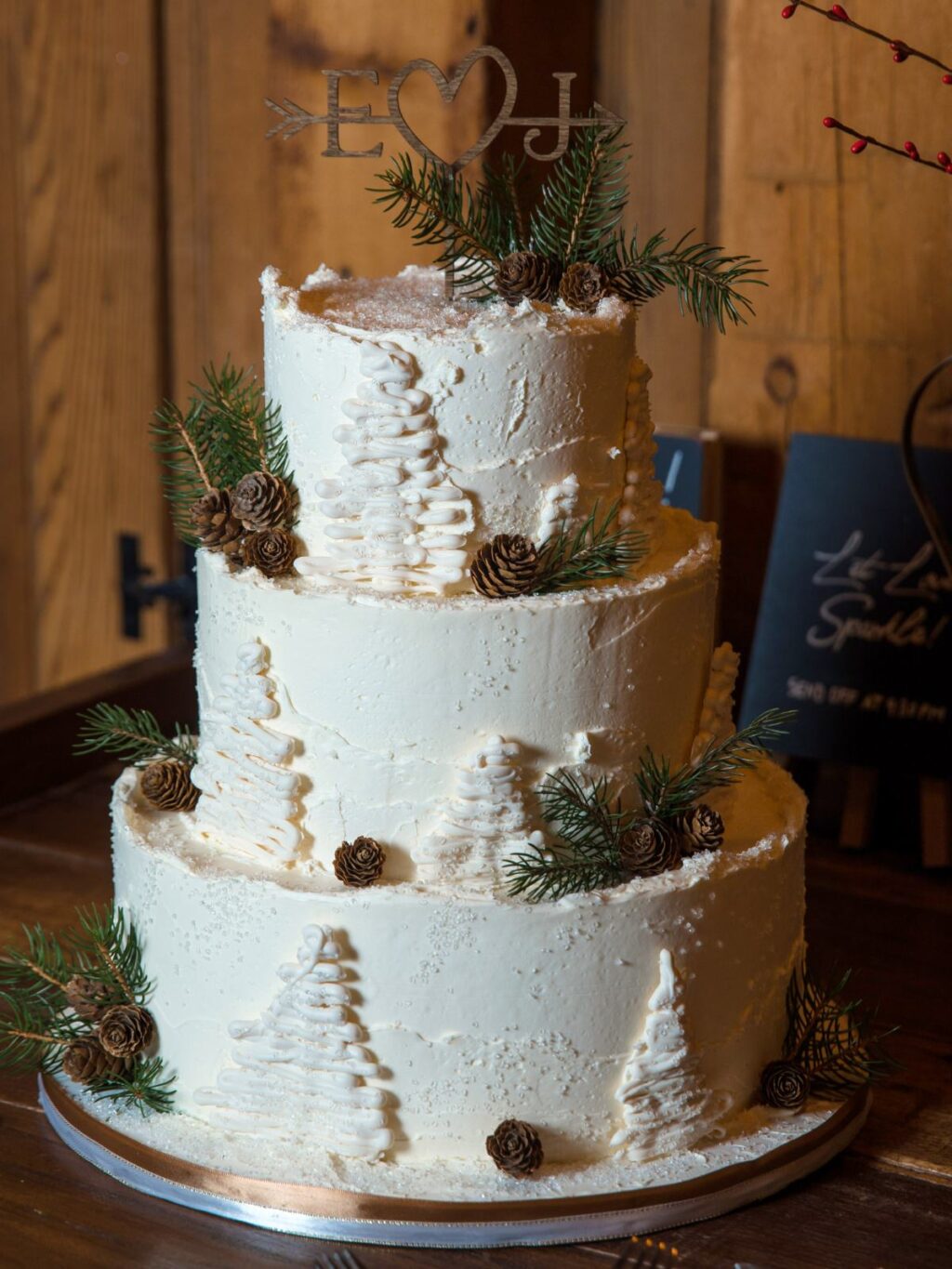 A cute winter wedding cake that is 3 tiers with frosting pine trees, sprigs of evergreen, and pinecones