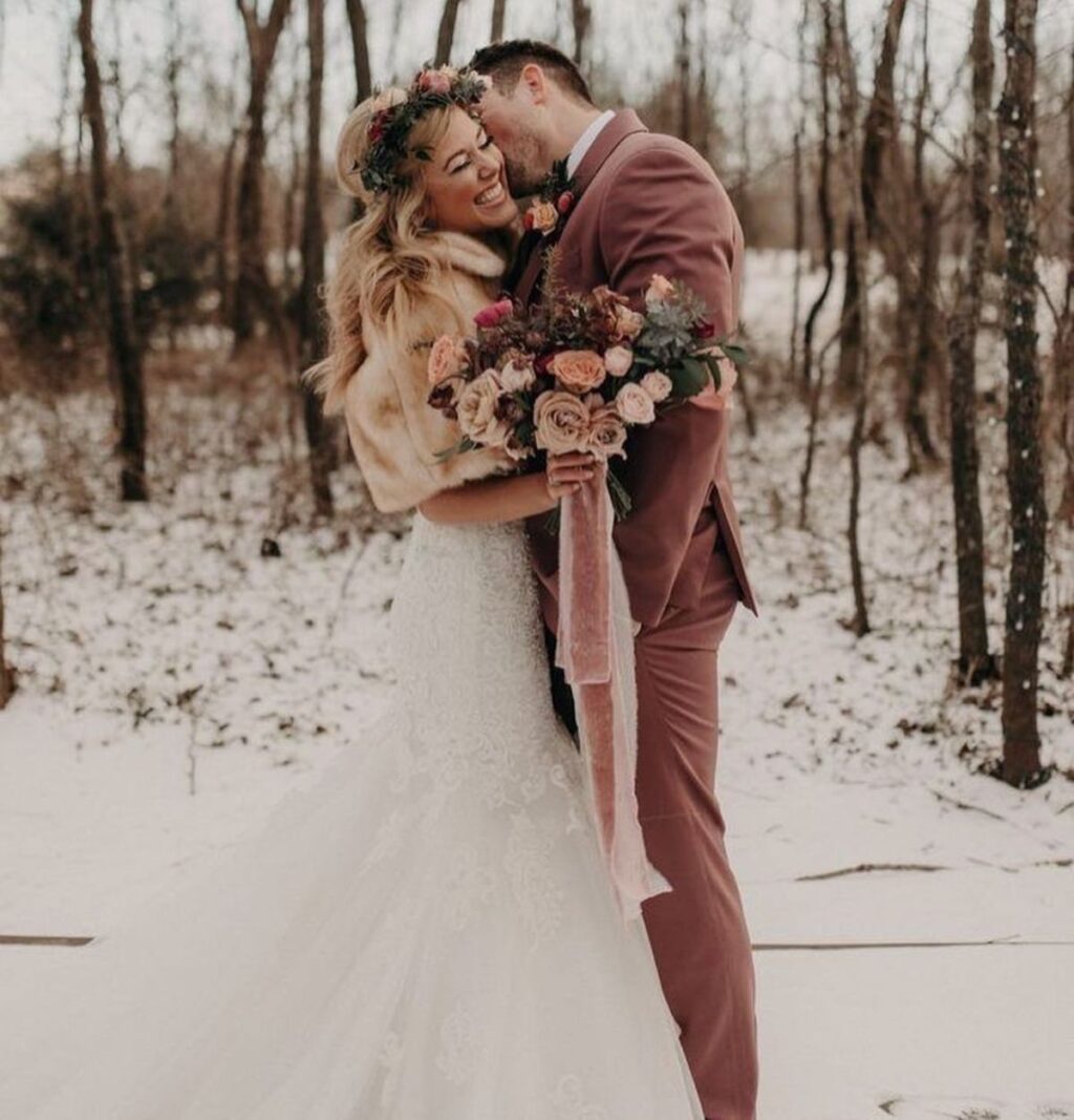A bride and groom posing in the snow. The groom is wearing a pink/red suit and the bride is wearing a dress with a winter faux fur wrap.