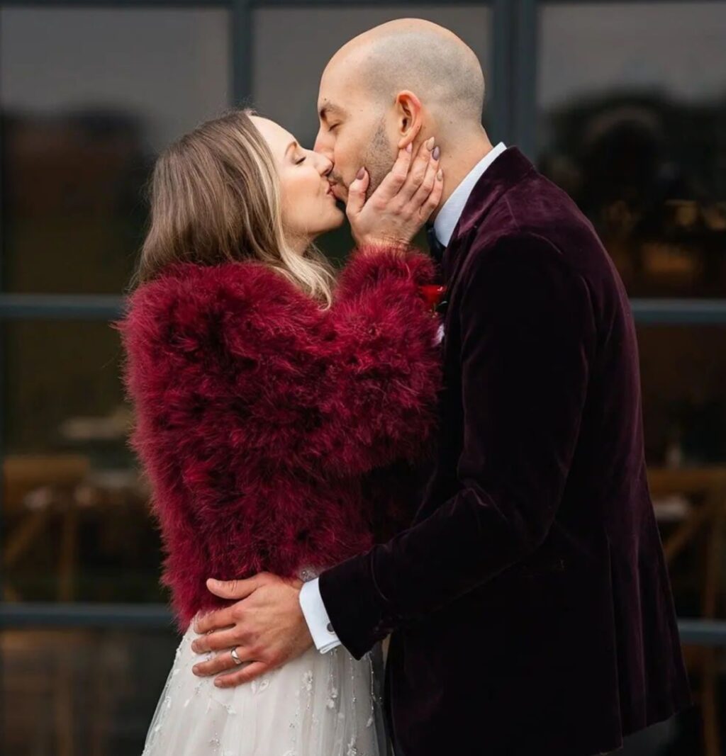 A bride and groom kissing. The bride is a wearing her white dress with a deep red faux fur wrap to stay warm