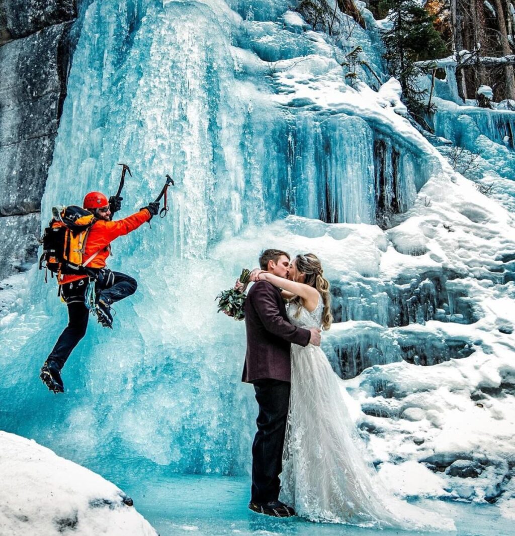 A bride and groom are kissing as an ice climber in a red jacket looks at them as he climbs up the frozen waterfall in the background