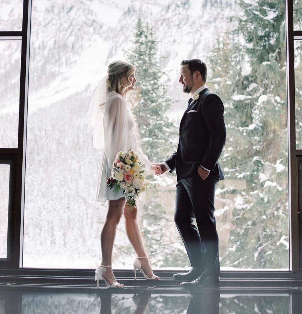 A couple posing in front of a window in the winter. The background is a snowy scene with snow covered trees and mountains