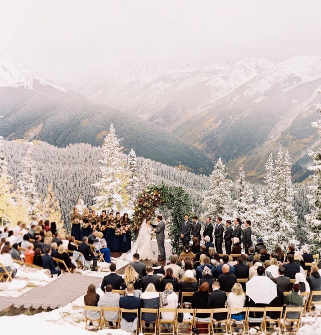 A beautiful outdoor winter wedding ceremony taking place in the mountains. The photo is taken from above and the guests are sitting in chairs and the bride and groom are getting married with snow covered trees in the background
