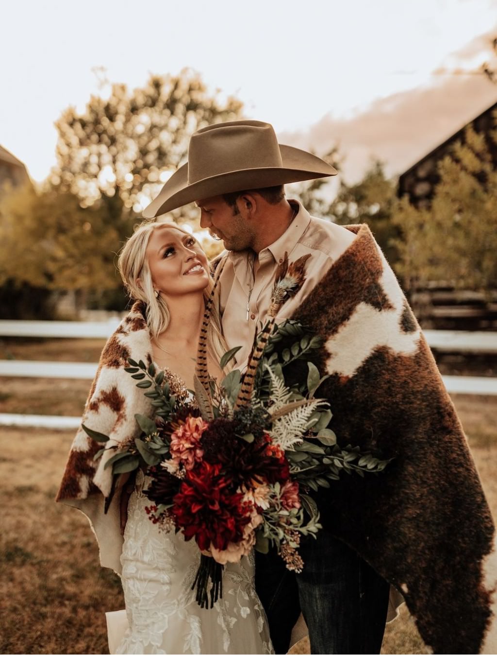 A rustic wedding photo that shows a bride and groom looking at each other and smiling. The groom is wearing a cow hide blanket and a cowboy hat