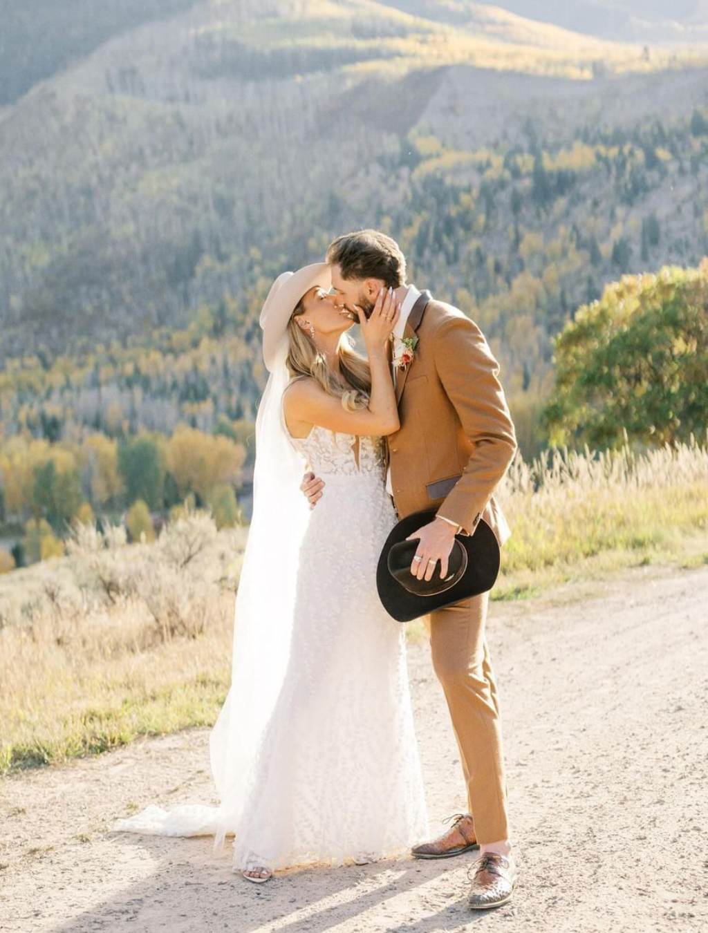 A romantic wedding photo that shows a bride and groom kissing on a dirt road. The groom has his cowboy hat removed and is holding it by his side.