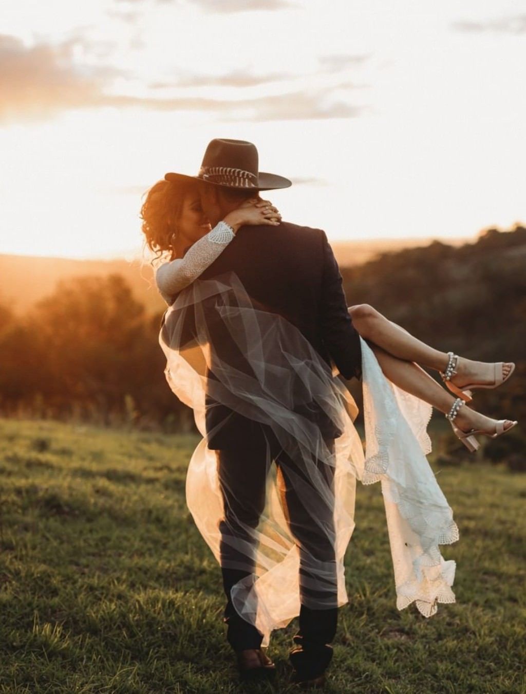 A beautiful wedding photo that shows a groom in a cowboy hat carrying the bride. They are walking away from the camera at sunset. It is a very romantic photo. This is for the blog post called rustic wedding photography ideas.