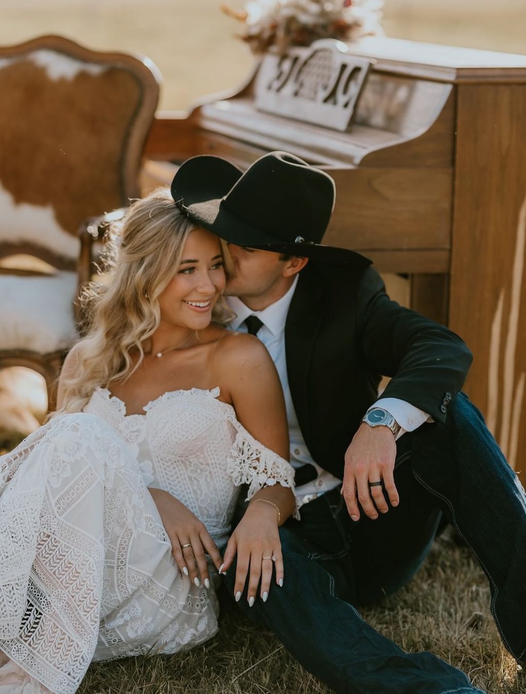 A bride and groom sitting in a field next to an antique piano. The groom is wearing a rustic cowboy hat