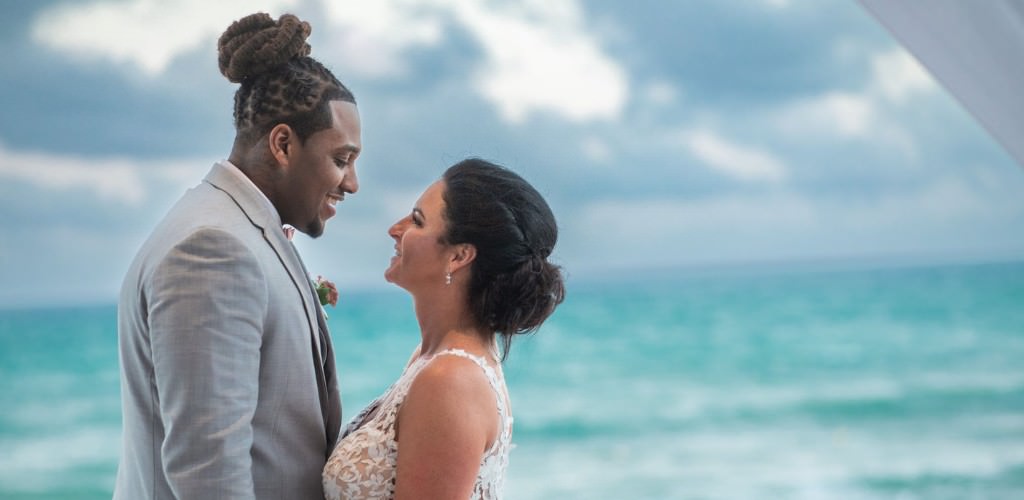 A bride and groom at the beach looking at each other. The bright blue ocean is behind them