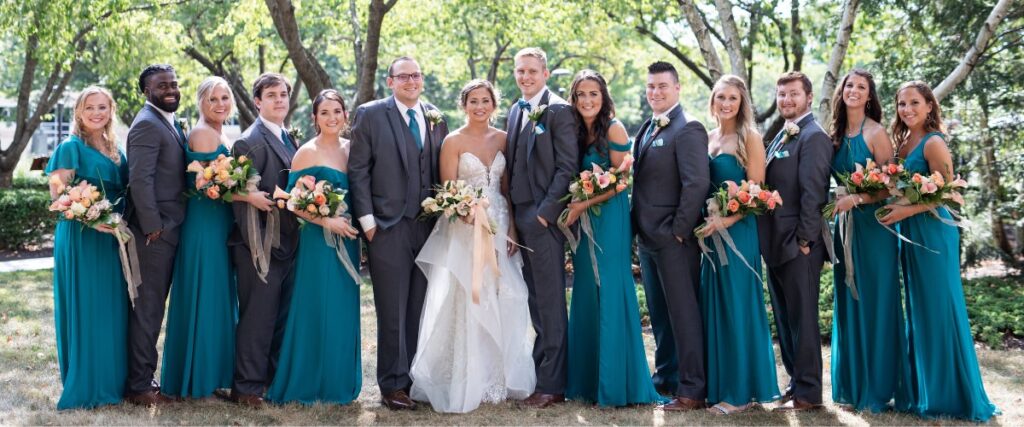 A wedding party posing for photos outside with trees behind them. they are wearing teal bridesmaid dresses and gray suits