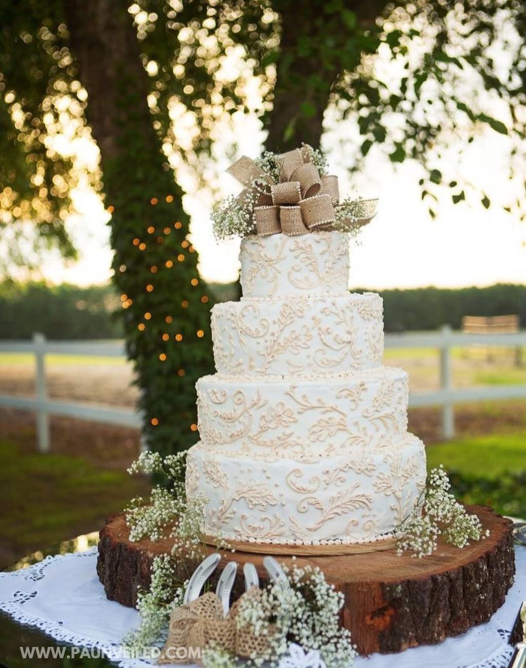 A 4-tier white wedding cake with a rustic feel to it. The cake sits on a tree log base and has a burlap bow cake topper