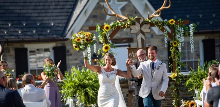 A bride and groom looking excited as they walk down the aisle after getting married. They ceremony arch behind them is decorated with deer antlers and sunflowers. This is the cover photo for the blog post titles rustic wedding ideas