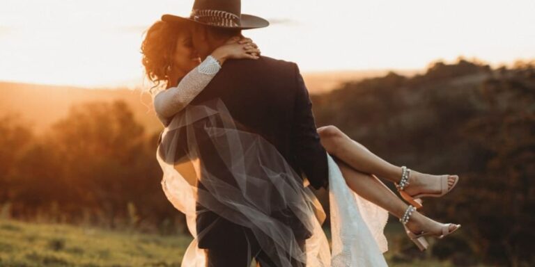 A groom with a cowboy hat carrying a bride in a field at sunset. This is the cover photo for the blog post called rustic wedding photography ideas