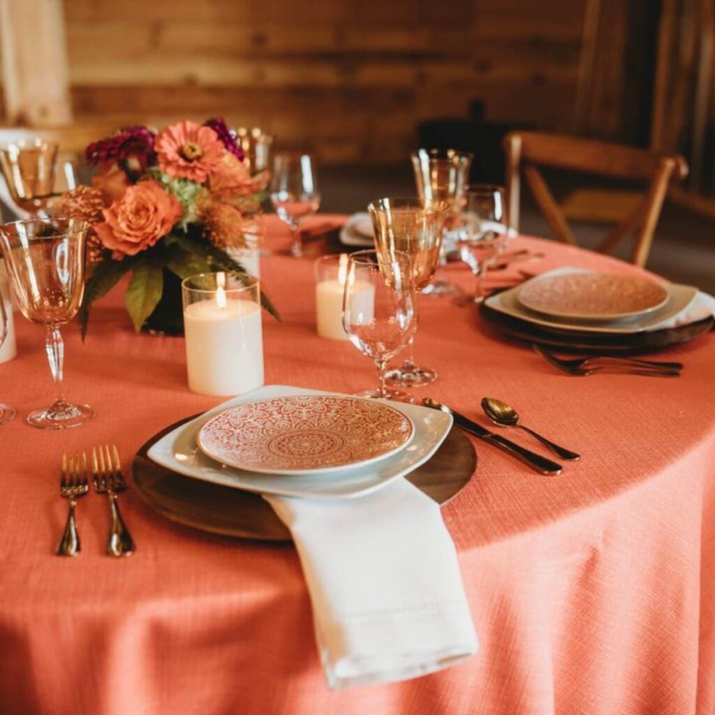 A closeup wedding reception detail photos that shows a table with an orange linen, a small centerpiece with orange flowers, and place settings and small candles.
