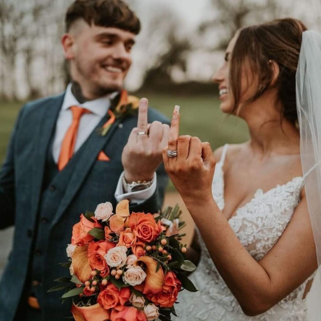A bride and groom holding up their ring fingers with their new wedding rings. The groom is wearing a dark suit with an orange tie. The bride is wearing a white dress and holding a bridal bouquet with a pretty orange color scheme