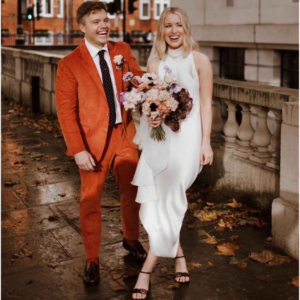 A bride and groom posing. The bride is in a white dress and the groom is wearing an orange suit,