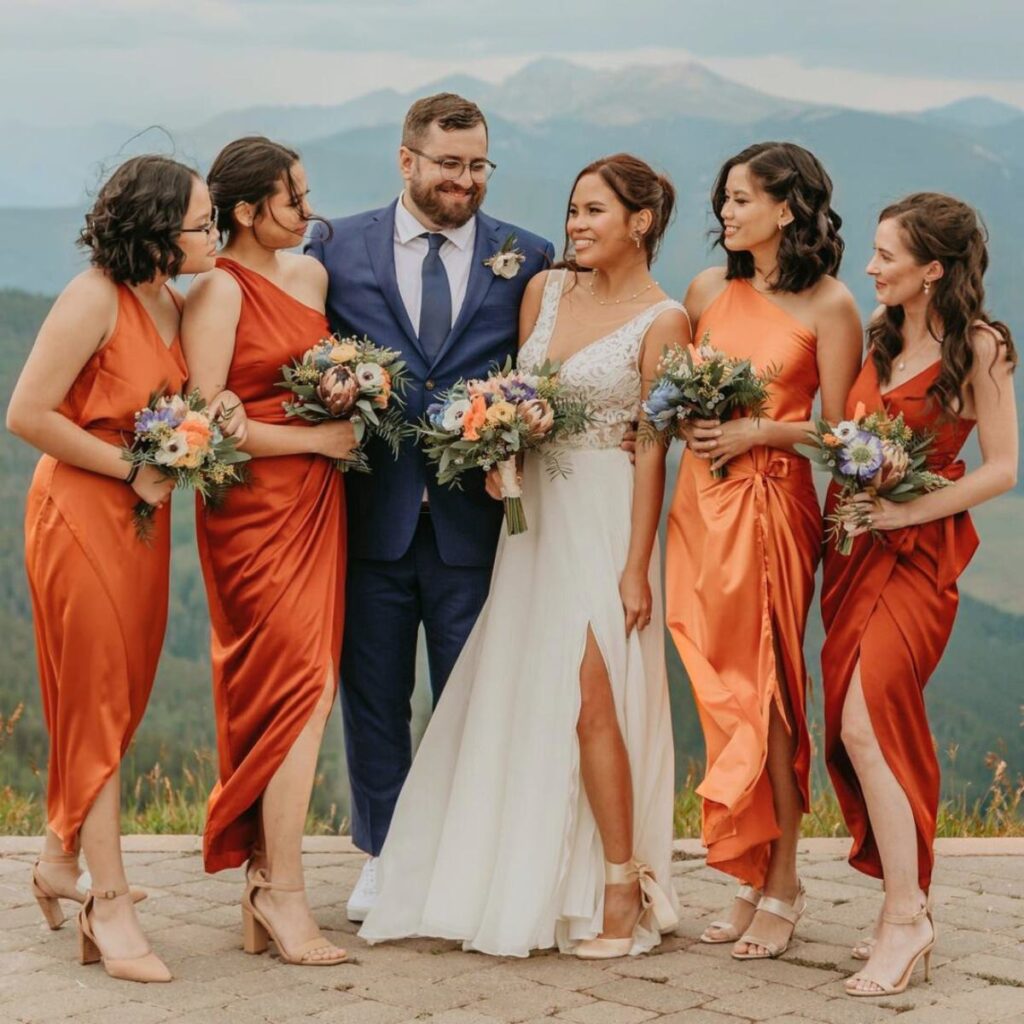 A groom in a gray suit with a bride in a white dress and four bridesmaids in orange dresses.