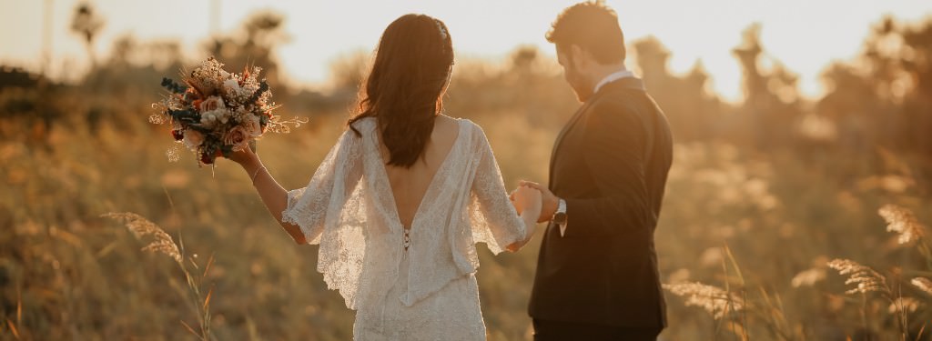 A bride and groom walking in a field at sunset