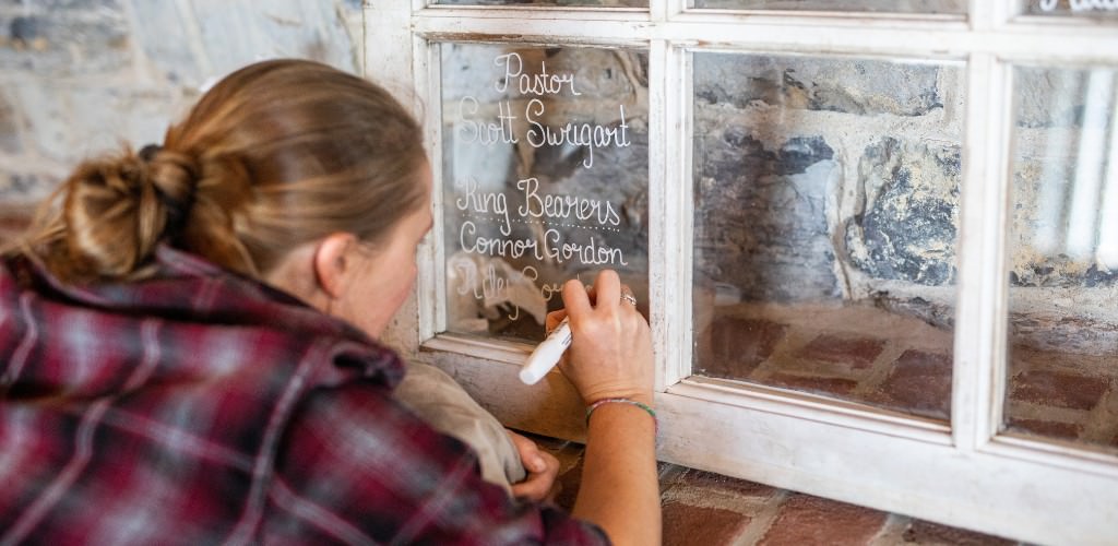 A bridesmaid writing wedding details in an old window pane to be displayed at the wedding.