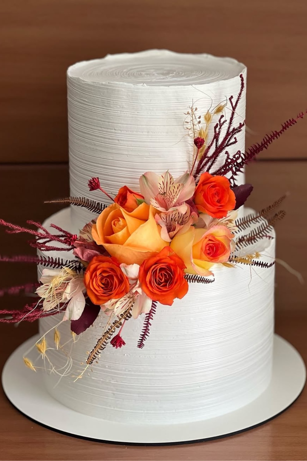 A pretty fall inspired white wedding cake with a 2-tier design. topped with fall flowers in oranges, reds, and yellows