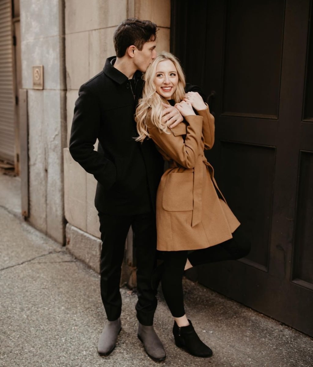 A couple posing in the city during winter. He is wearing all black and she is wearing black pants with a stylish brown coat or jacket.