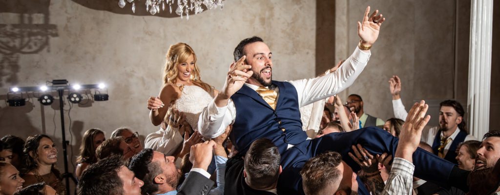 A bride and groom being lifted in the air at their wedding reception as they dance on the dance floor