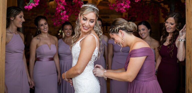 A bride having her wedding dress buttoned by her mom as her bridesmaids watch