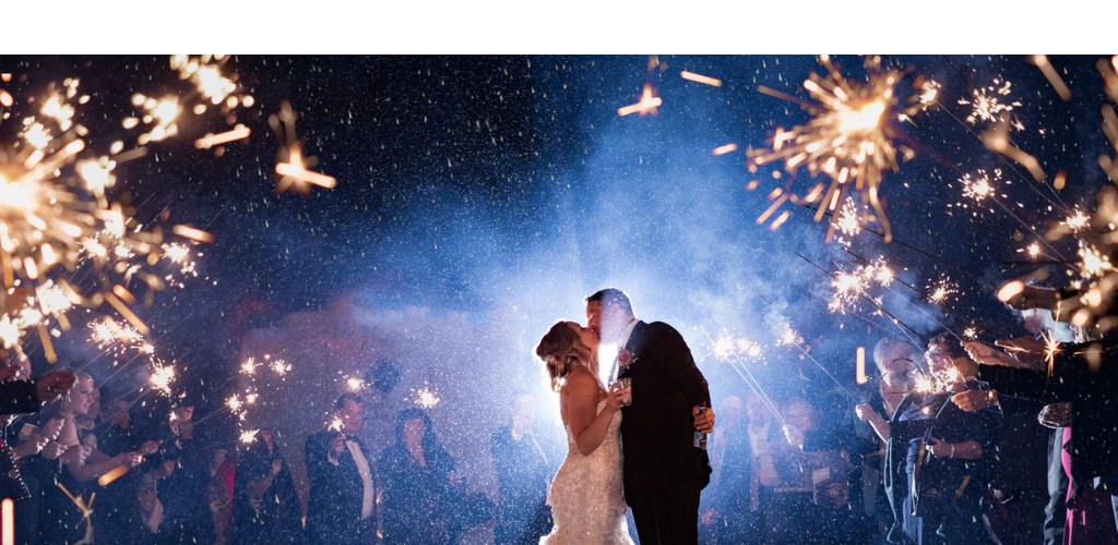 A bride and groom kissing at the end of their reflection. It is raining outside and all of the guests are holding sparklers.