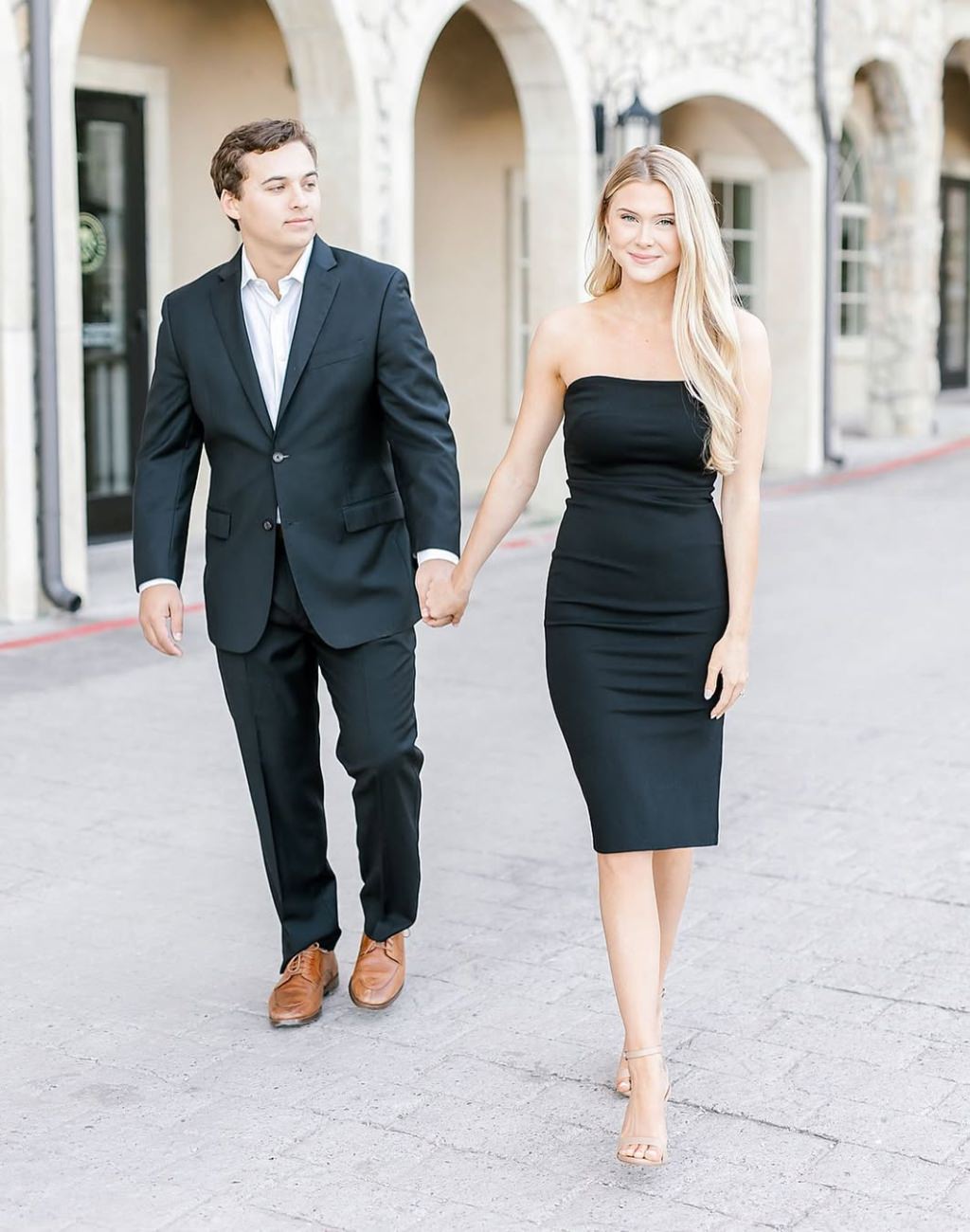 A couple posing for an engagement photos and the girl is a black dress and the guy is in a black suit with white shirt.