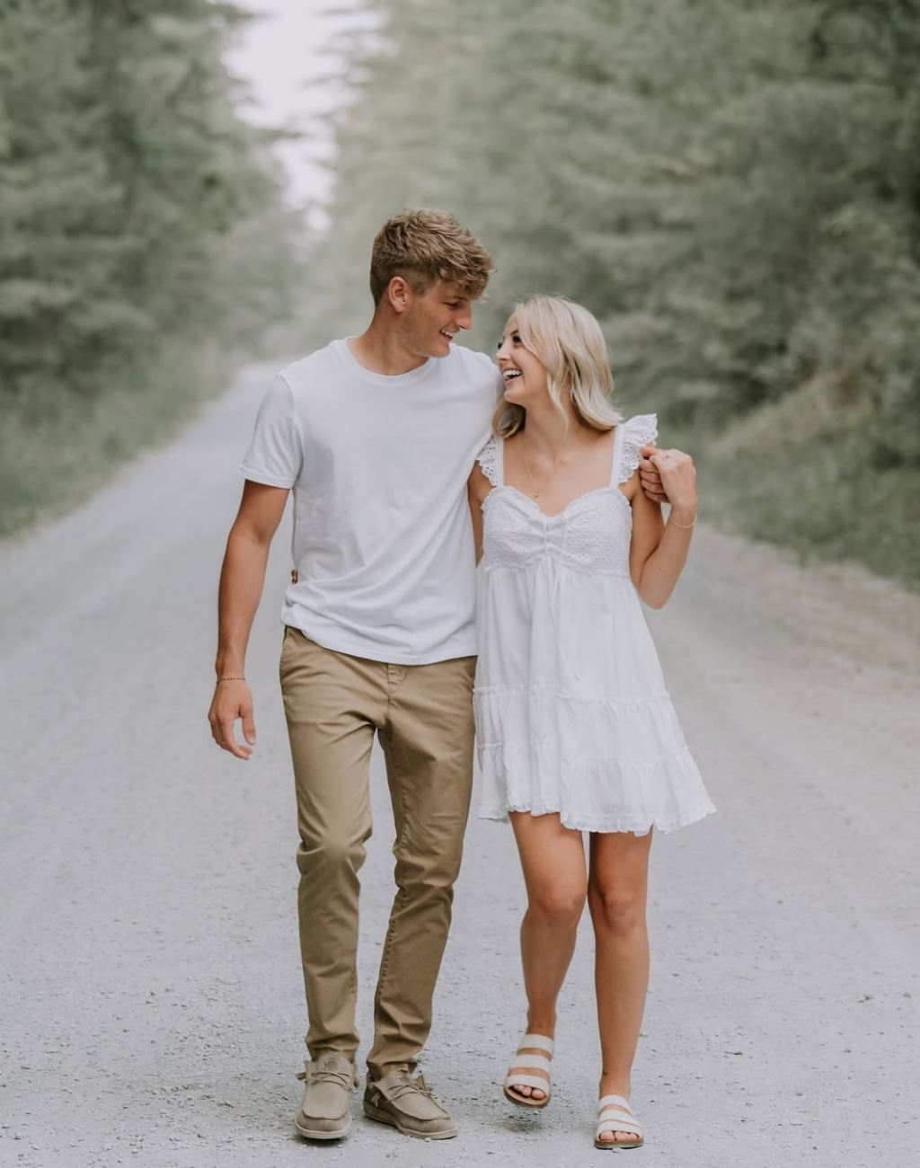 A cute couple walking in the middle of the road for their engagement pictures. They have their arms around each other and are looking at each other and smiling. The girl is in shot white sundress.