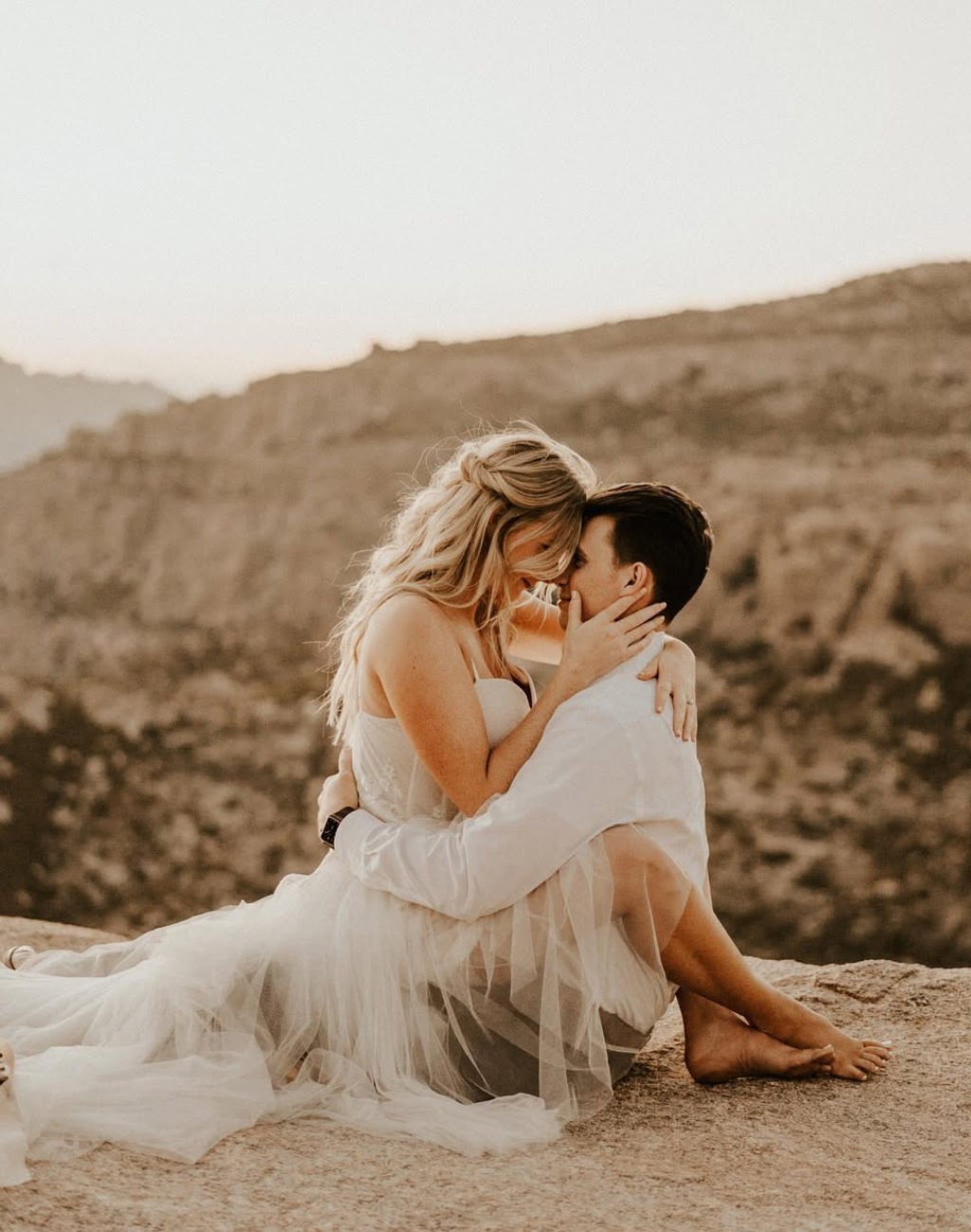 A romantic pose for this engagement photo that shows the couple closely holding each other, the girl sitting on the guy's lap and she is in a dress and barefoot.