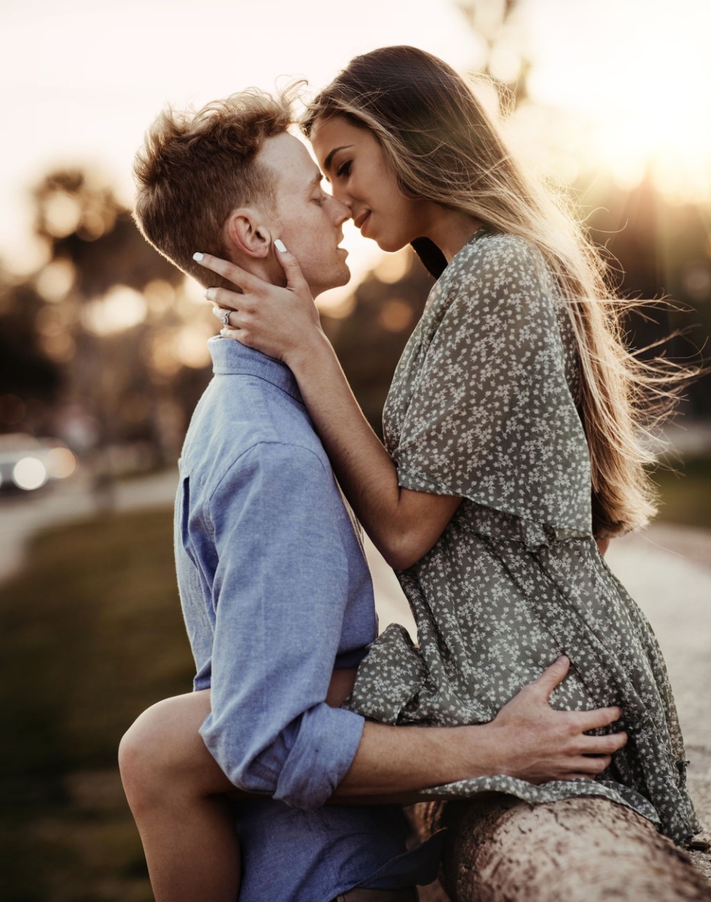 A romantic engagement photo at sunset with the girl sitting on a fence post as the guy leans in and kisses her.