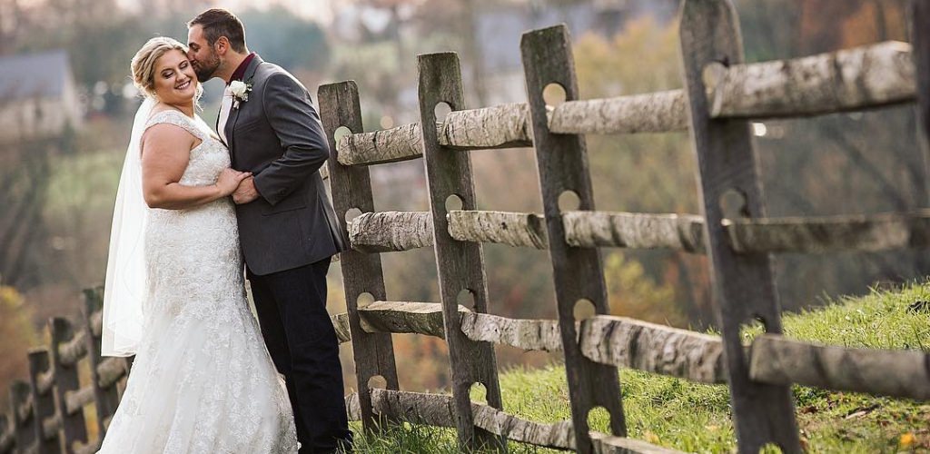 A bride and groom kissing near a wooden fenxr. This is for the blog post called how to create a wedding budget
