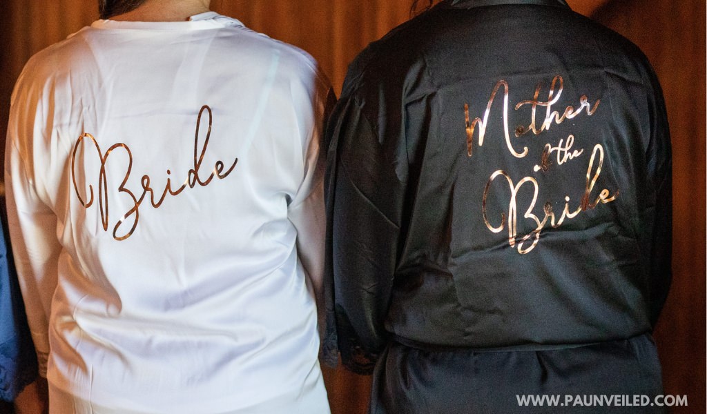 A mom and the mother of the robe wearing robes on the wedding day. The bride's robe says bride. And the mother of the bride's robe says mother of the bride.