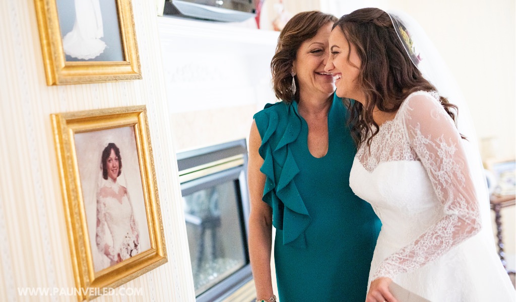 The bride and her mom looking at an old bridal portrait of the mom. The bride is wearing the mom's dress on her wedding day