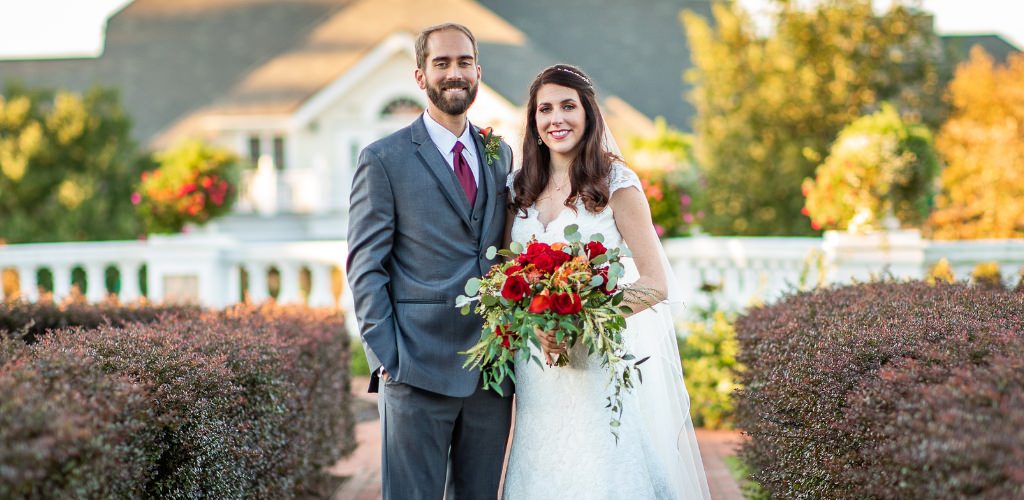 A bride and groom posing for the camera outdoors. The bride is holding a red bridal bouquet