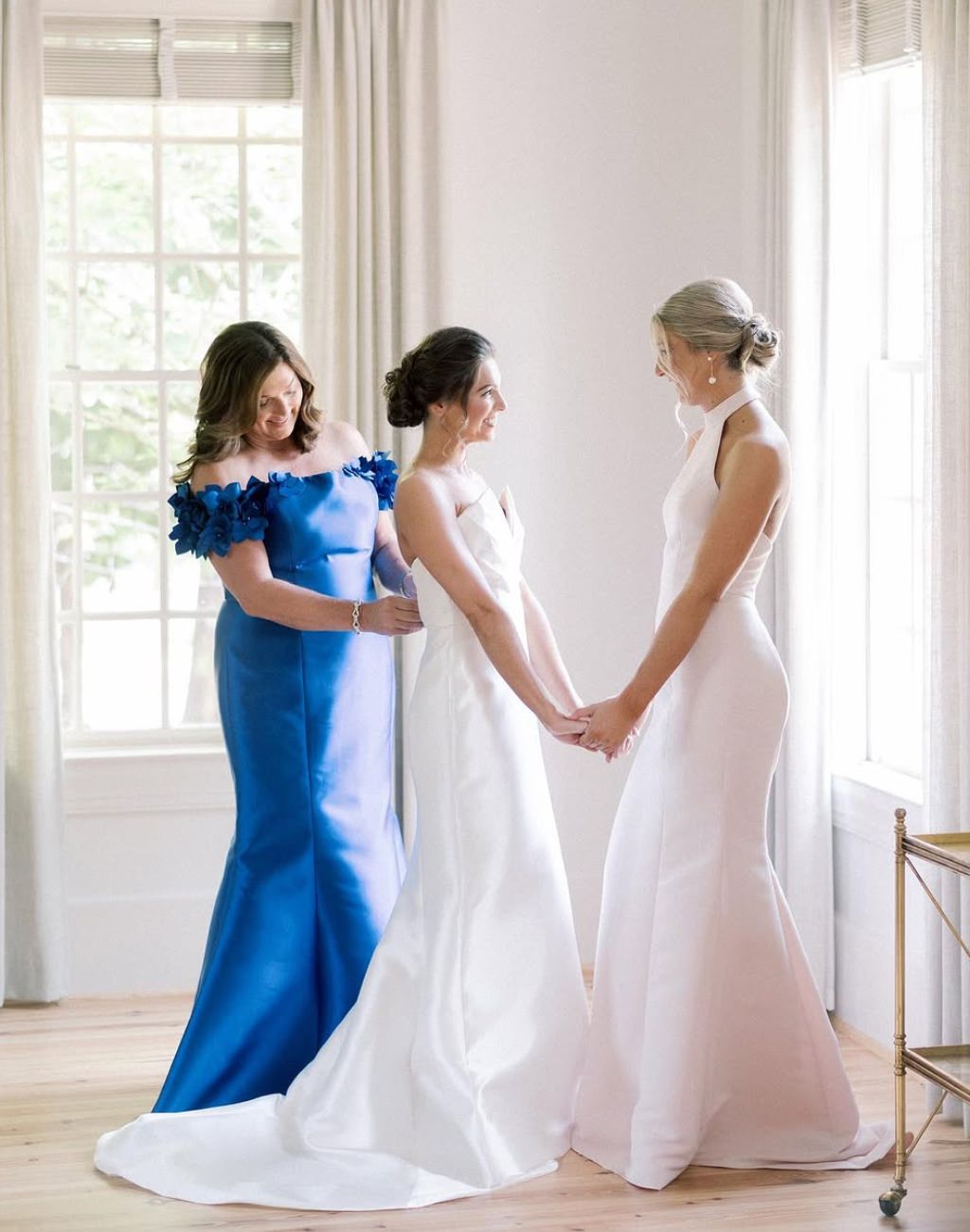 A mother of the bride wearing a bright blue dress zipping up the bride's dress as the bride's sister holds the bride's hands.