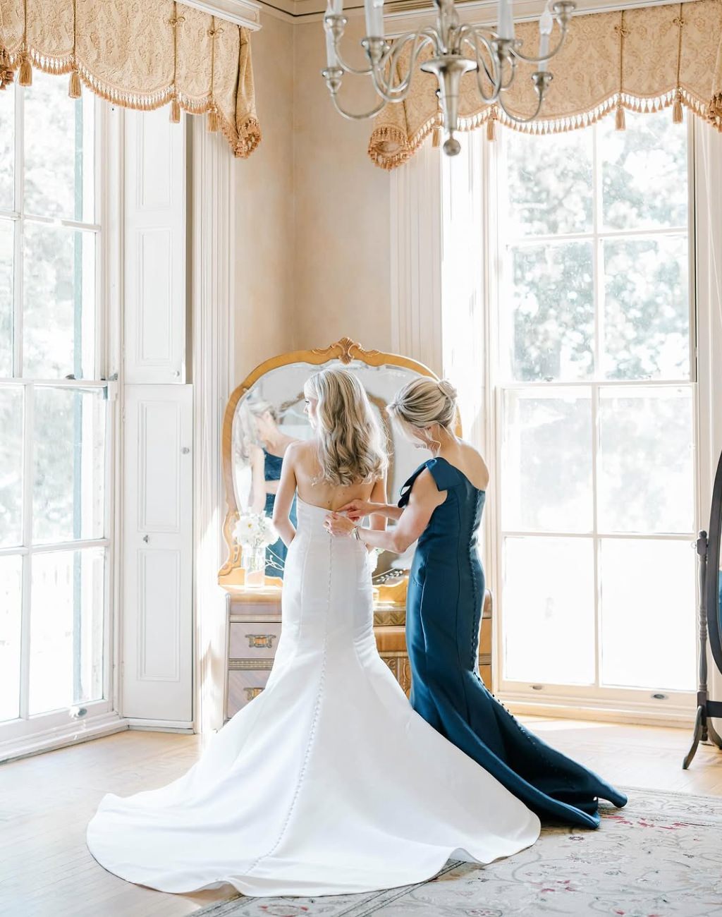 A bride's mom zipping up her daughter's wedding dress in a big room lit by 2 big windows
