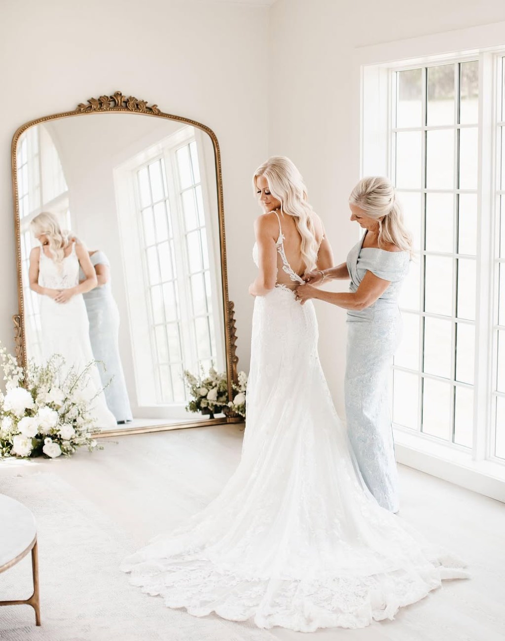 A bride's mom zipping up the bride's wedding dress in front of a big mirror