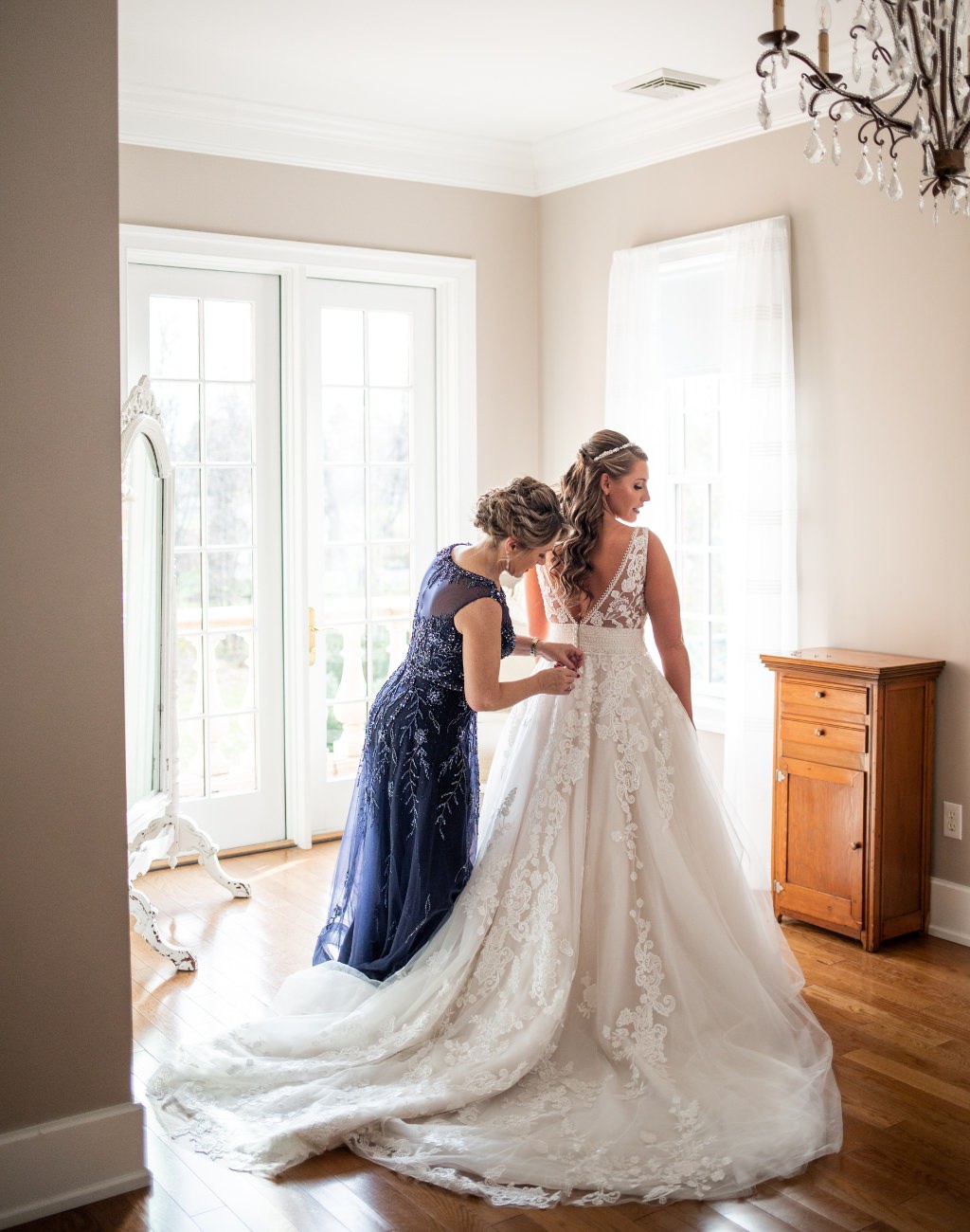 A bride's mom zipping up the bride's wedding dress in a window lit room